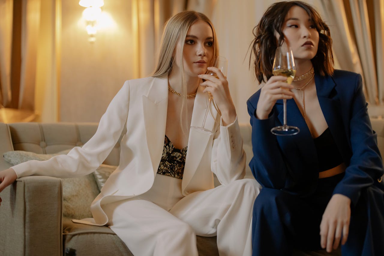 Two fashionable women in tailored suits enjoying a glass of wine indoors, exuding glamour and friendship.